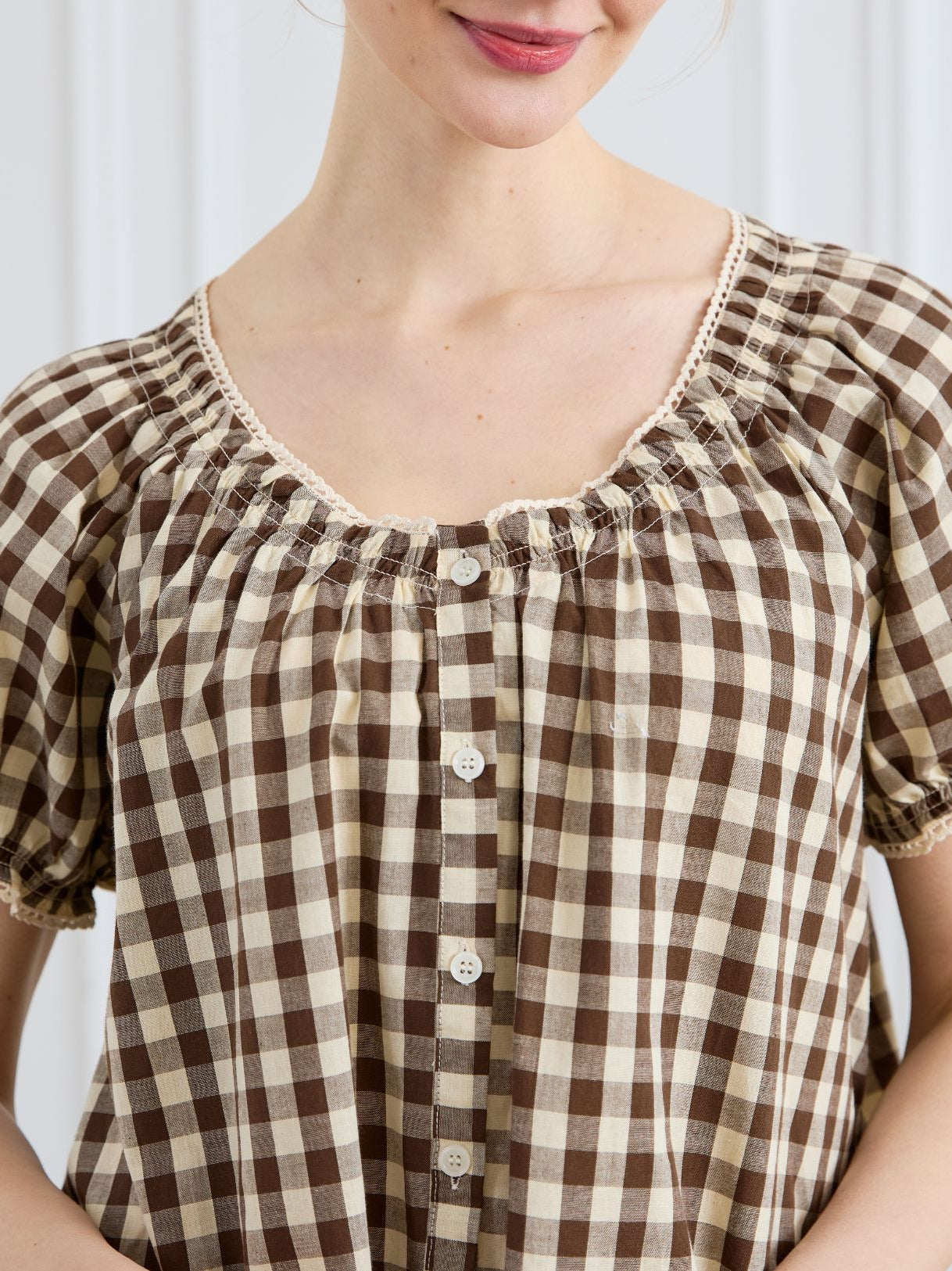 Close-up of a woman smiling, wearing a brown and cream gingham 100% cotton dress, featuring a gathered neckline with delicate lace trim and a button-down front.