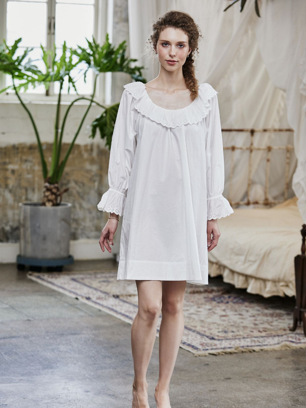 Full-length view of a woman wearing an oversized Renaissance cotton nightgown with ruffled sleeves and romantic lace trim in a vintage bedroom setting.