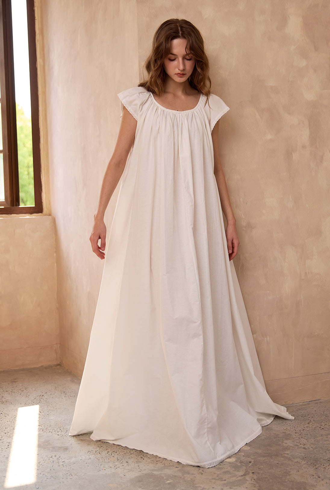 Sueasyretro ivory cotton Victorian nightgown near a window. Floor-length modest nightie with an elegant, roomy drape.