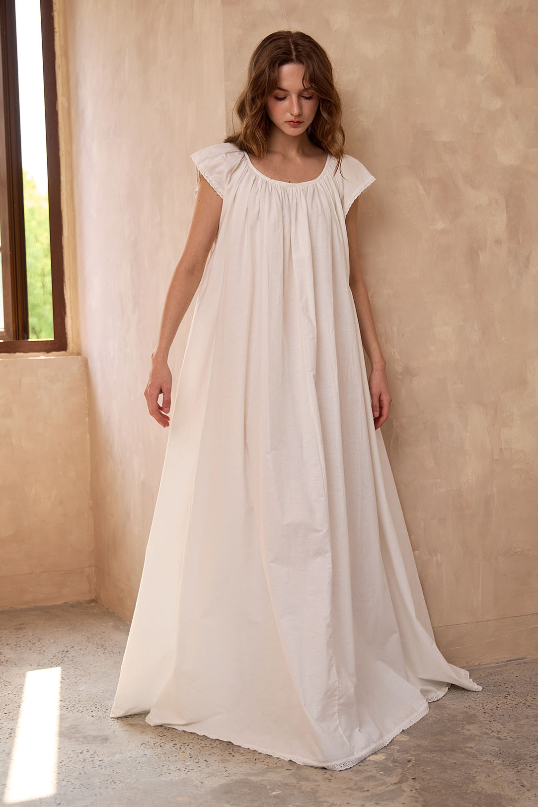 Sueasyretro ivory cotton Victorian nightgown near a window. Floor-length modest nightie with an elegant, roomy drape.