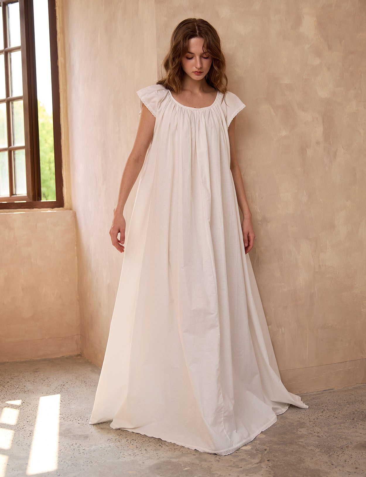 Sueasyretro ivory cotton Victorian nightgown near a window. Floor-length modest nightie with an elegant, roomy drape.