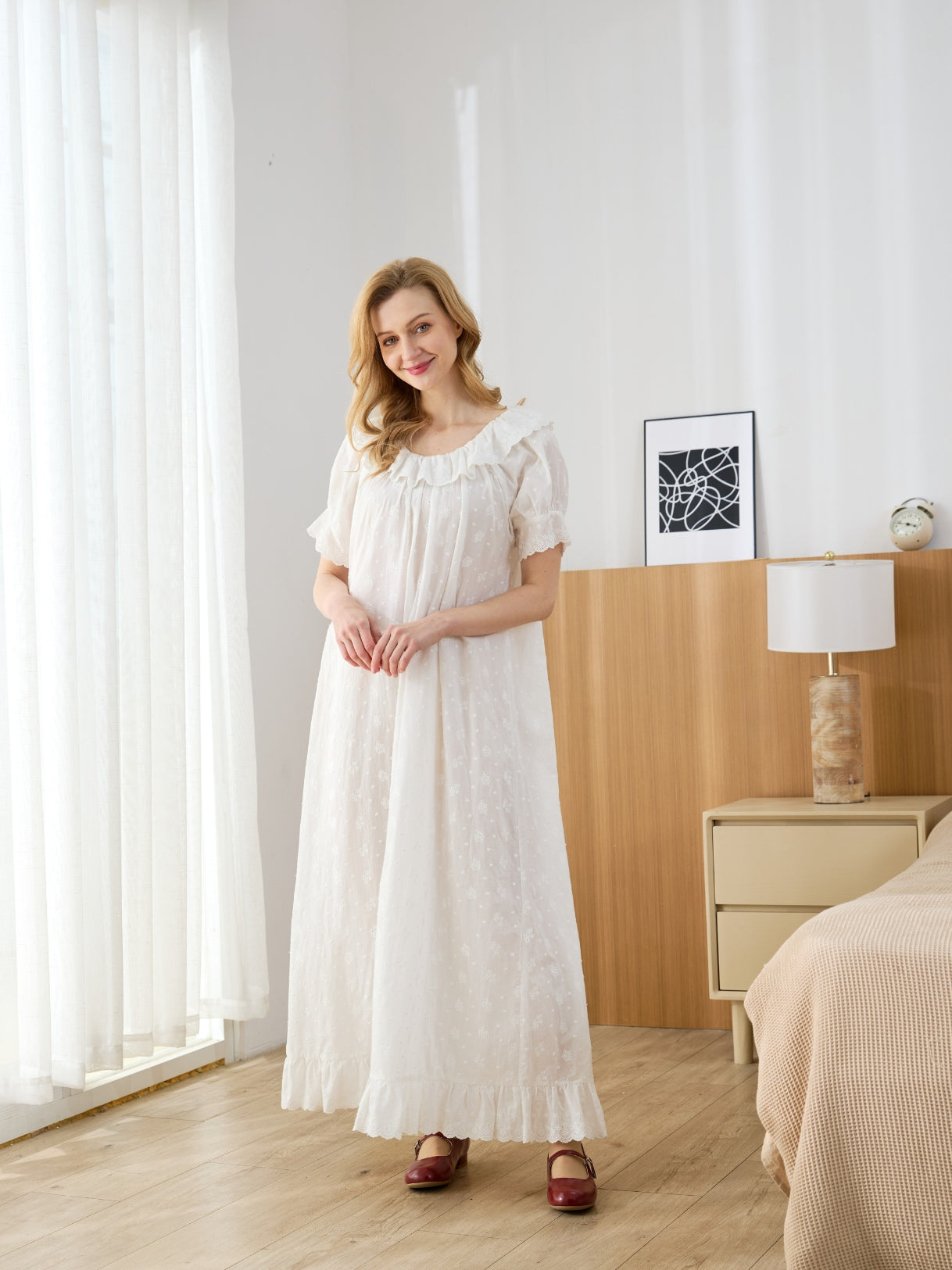 Full-length front view of a woman wearing an off-white Victorian cotton nightgown with dotted floral embroidery in a bright bedroom.