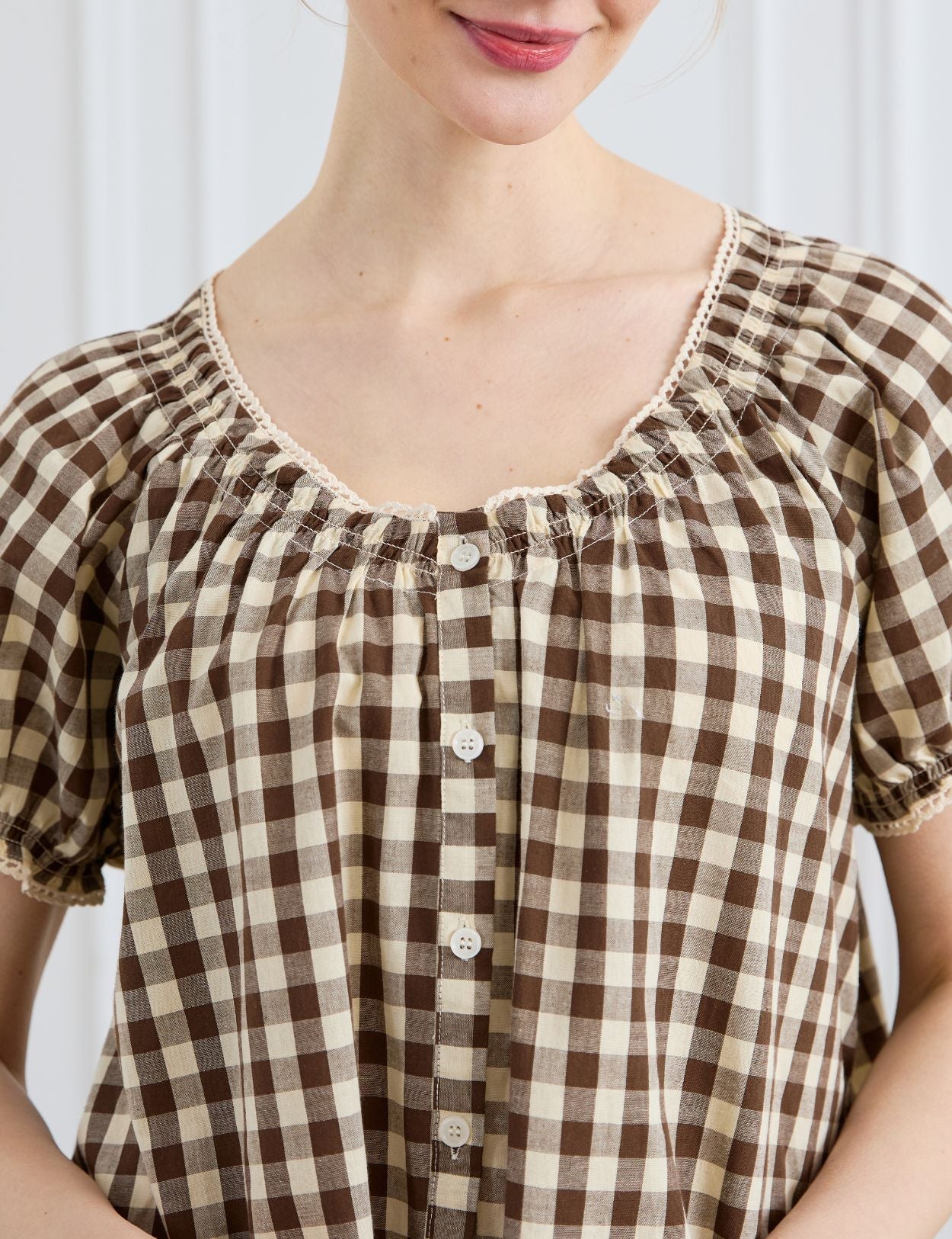 Close-up of a woman smiling, wearing a brown and cream gingham 100% cotton dress, featuring a gathered neckline with delicate lace trim and a button-down front.