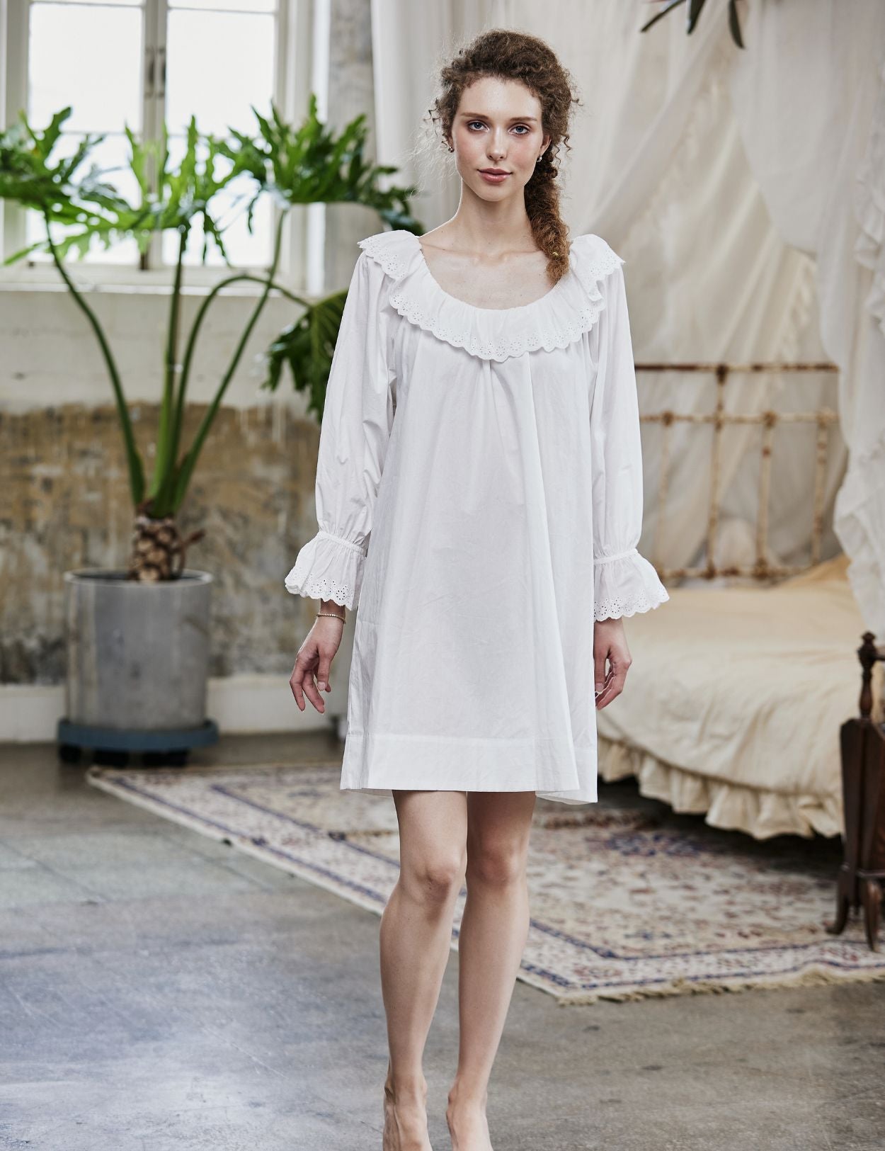 Full-length view of a woman wearing an oversized Renaissance cotton nightgown with ruffled sleeves and romantic lace trim in a vintage bedroom setting.