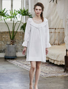 Full-length view of a woman wearing an oversized Renaissance cotton nightgown with ruffled sleeves and romantic lace trim in a vintage bedroom setting.