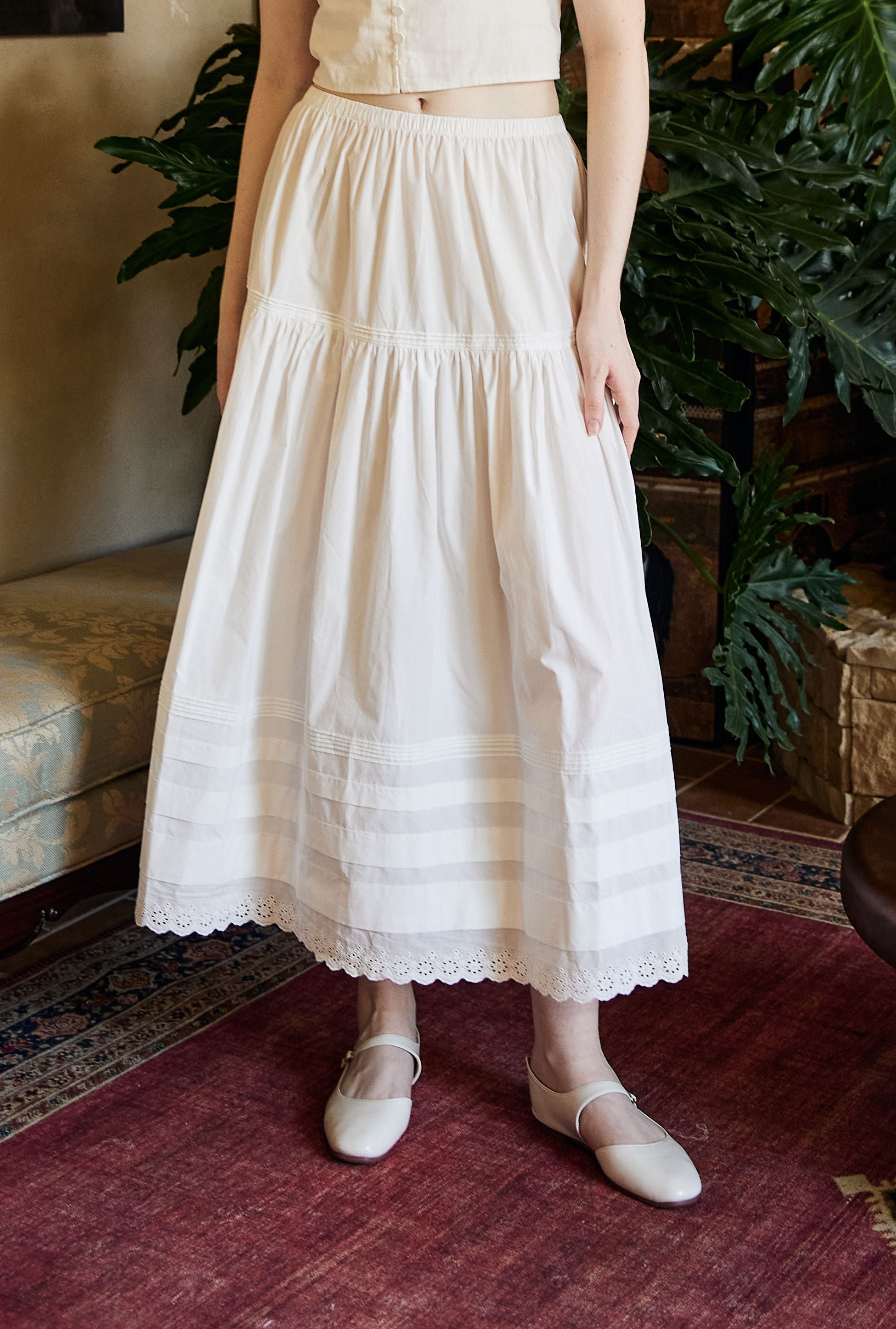 Close-up of a pure cotton Victorian-style maxi skirt, showcasing horizontal tiered pintucks and a romantic fan-shaped hemline with delicate floral eyelet embroidery.