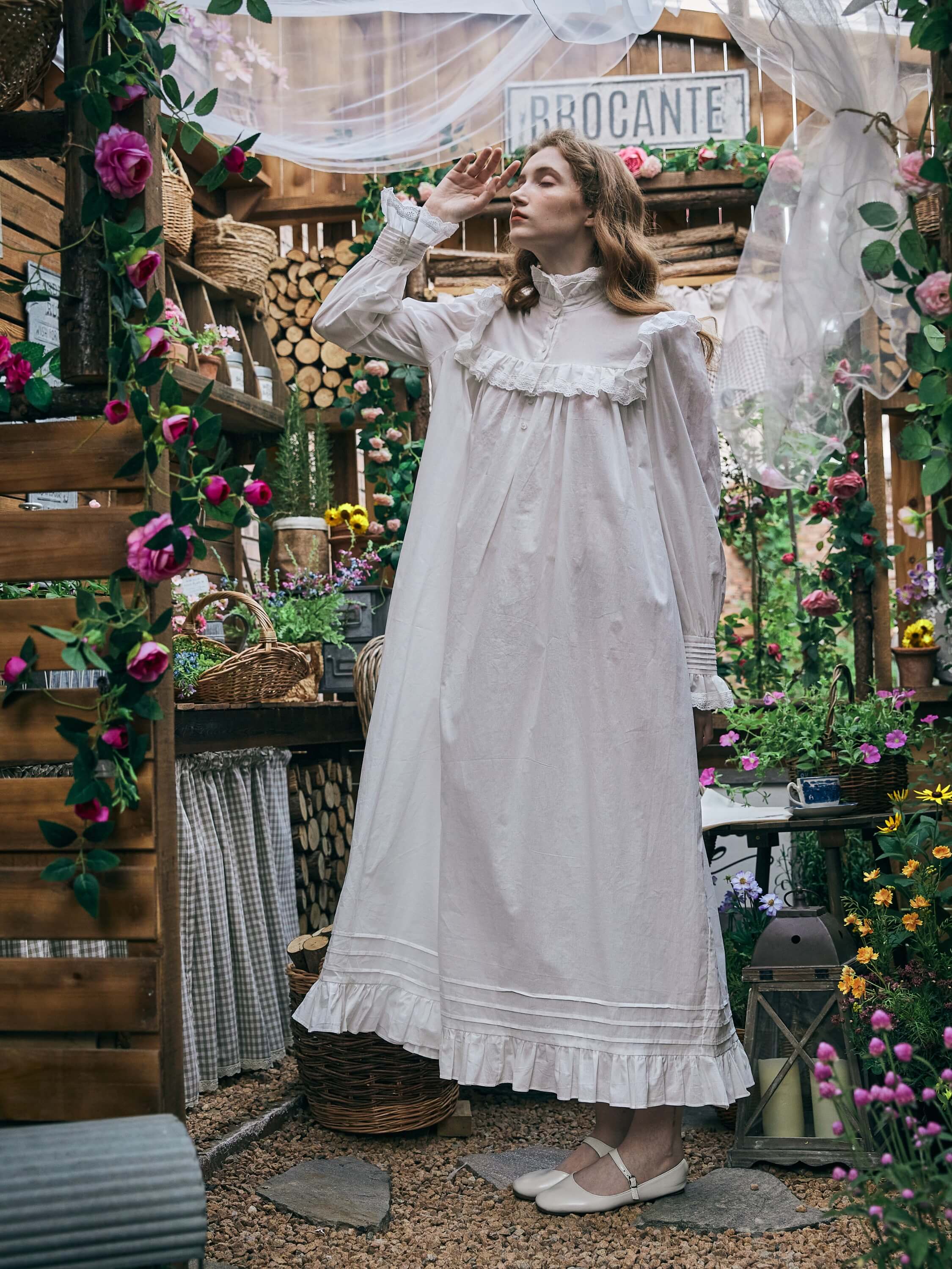 Woman standing in a rustic greenhouse garden, wearing the white cotton Edwardian-style nightgown. She is looking upwards with one hand raised near her face, showcasing the long bishop sleeves and flowing silhouette.