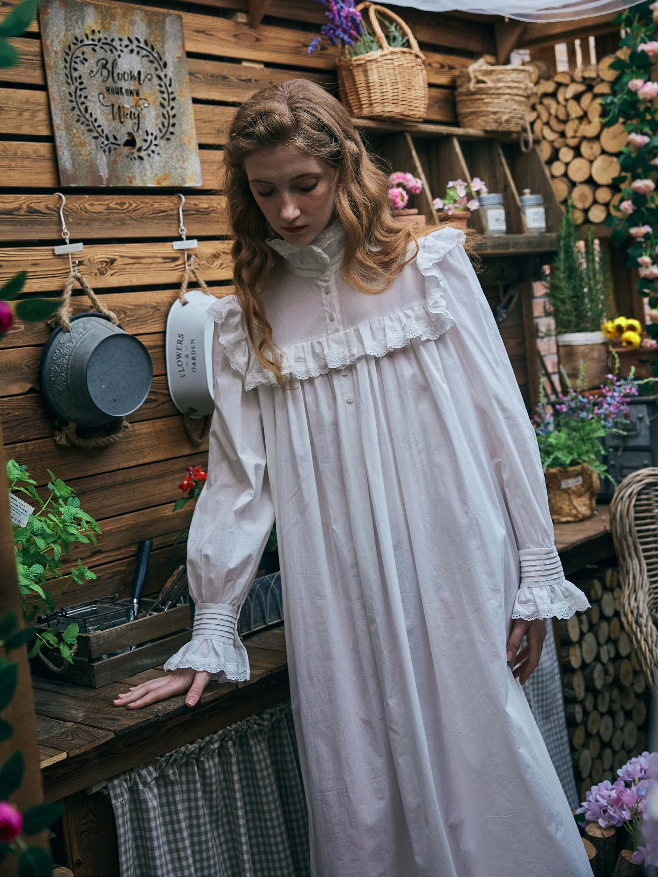 Woman wearing the Ophelia white Edwardian cotton nightgown standing in a rustic shed, looking down while resting her hand on a wooden table. The image highlights the ruffled yoke and long bishop sleeves.