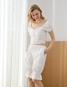 Close-up of a woman wearing a rustic Edwardian-style white cotton outfit featuring a button-up puff-sleeve top and elastic waist bloomers with pockets.