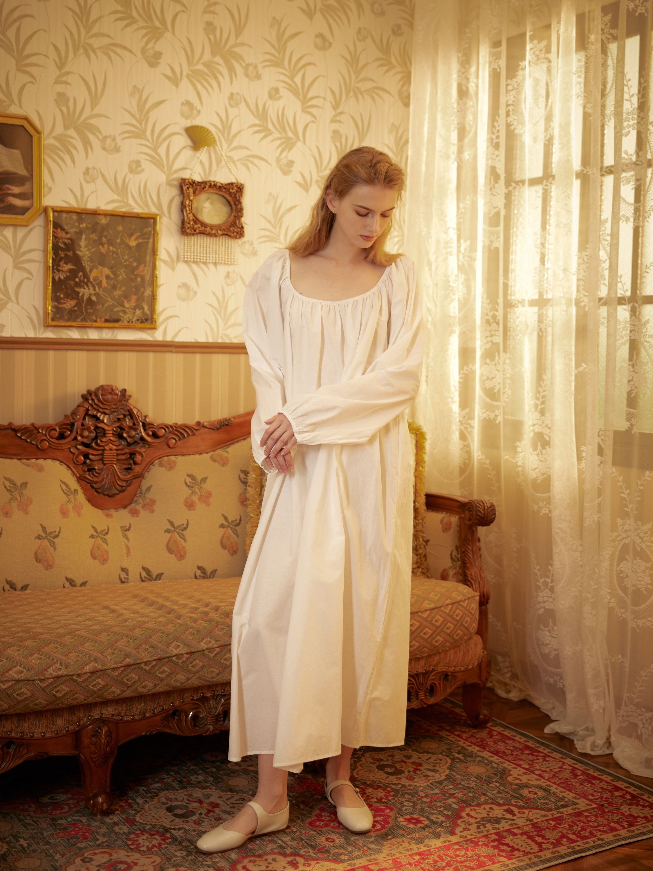 Woman standing in a vintage room wearing a white Victorian cotton nightgown with a square neck and long sleeves.