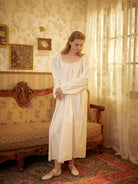 Woman standing in a vintage room wearing a white Victorian cotton nightgown with a square neck and long sleeves.