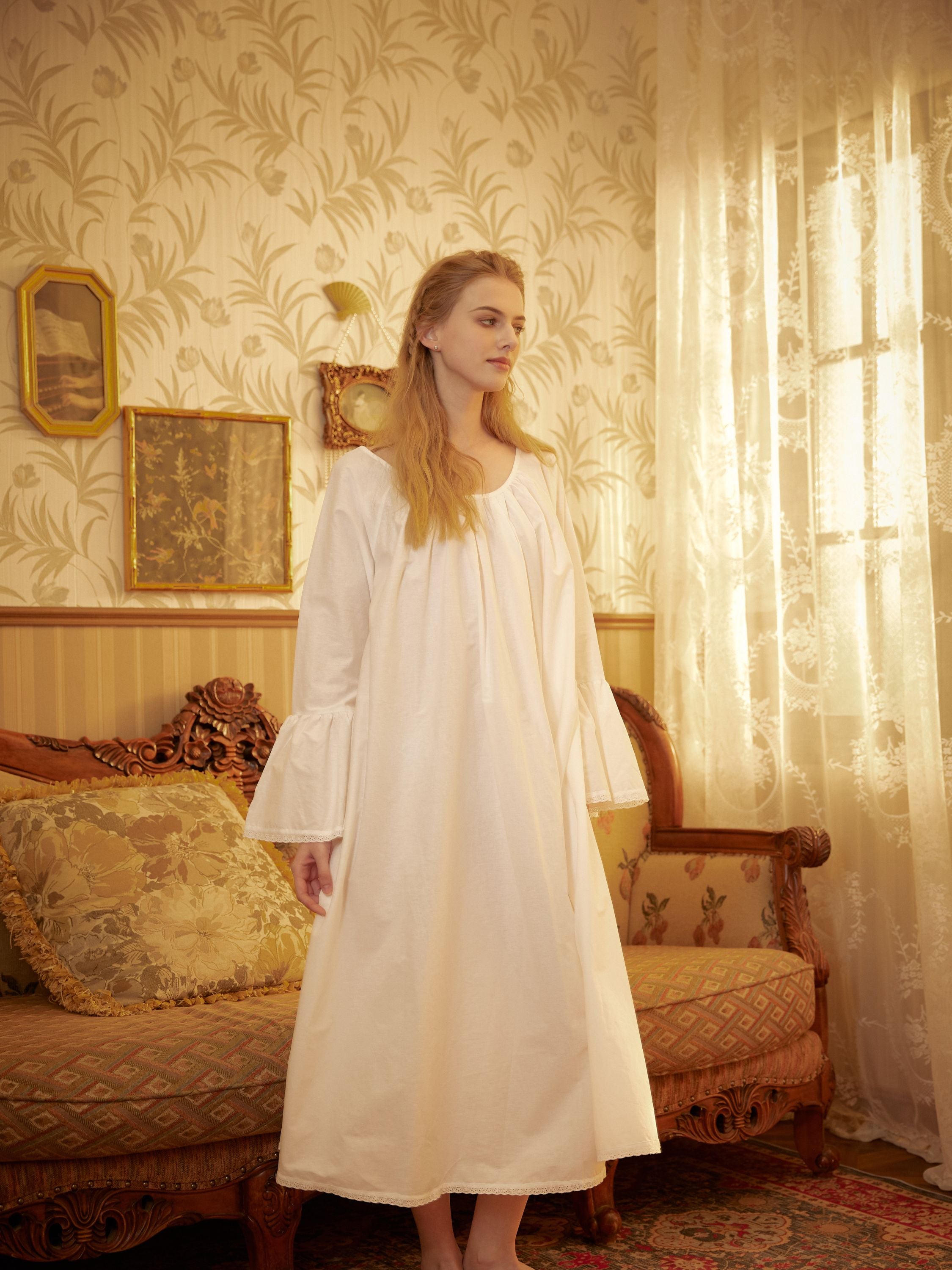 Model standing in a vintage Victorian parlor wearing a long white cotton nightgown with ruffled sleeves, standing next to an ornate antique sofa. Natural light from a window highlights the floral wallpaper background.