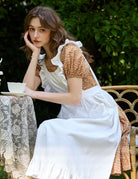 Woman seated wearing ivory Victorian cotton pinafore apron with ruffle trim