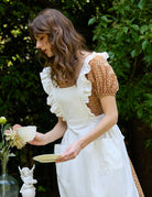 Woman wearing ivory cotton pinafore apron with ruffle trim, hands in front pockets